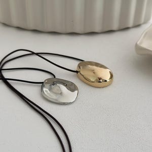 May include: Two necklaces with a black cord and a silver and gold pendant. The pendants are both oval shaped and have a smooth, shiny finish.