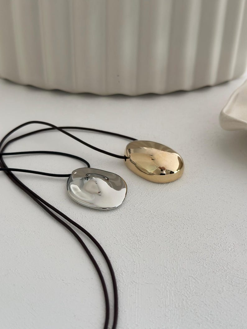 May include: Two necklaces with a black cord and a silver and gold pendant. The pendants are both oval shaped and have a smooth, shiny finish.