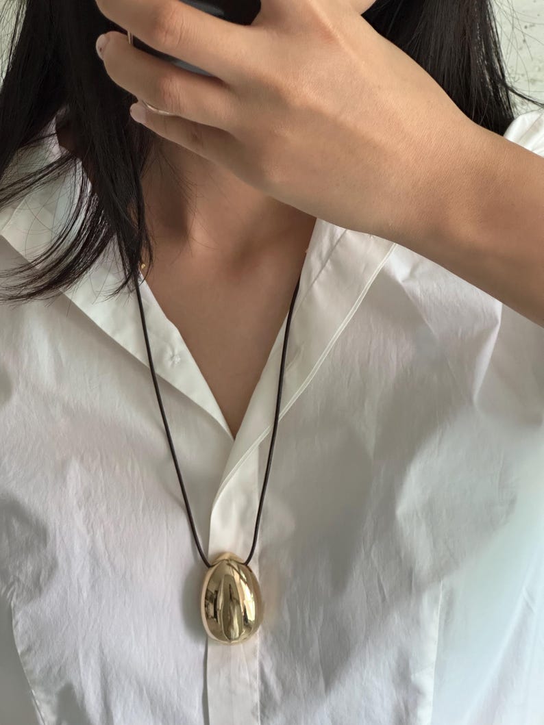 May include: A gold pendant necklace with a smooth, rounded, egg-shaped charm. The necklace is on a black cord.