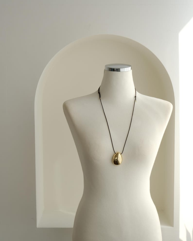 May include: A gold teardrop pendant necklace on a black cord, displayed on a white mannequin.