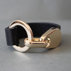 May include: A black leather bracelet with a gold-colored clasp. The bracelet features a wide, textured leather band and a large, circular gold-tone clasp. The clasp is connected to a gold-colored rectangular piece. The bracelet is a stylish accessory.