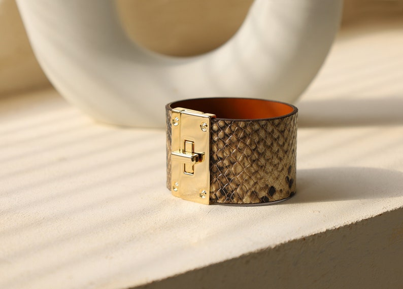 K&ouml;nnte beinhalten: Ein goldfarbenes und braunes Lederarmband mit Schlangenhautmuster und einem goldenen Verschluss.
