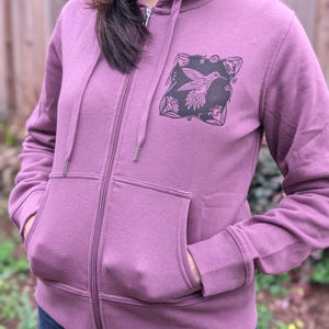 May include: A mauve zip-up hoodie with a black and white hummingbird design on the chest. The hoodie has a drawstring hood, front pockets, and ribbed cuffs. The zipper is silver.
