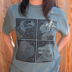 May include: A sage green short-sleeve t-shirt featuring a black graphic print divided into four rectangular panels. Each panel displays a detailed line drawing of a reptile or sea creature accompanied by handwritten-style scientific text.