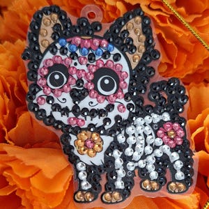 Puede incluir: Un dije decorativo con forma de perro, adornado con pedrería negra, blanca, rosa y transparente. El perro presenta un diseño de calavera de azúcar en su rostro y una flor en su costado, sobre un fondo naranja.