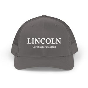 May include: A gray trucker hat with the word "LINCOLN" in white block letters above "Cornhuskers football." The hat has a mesh back and a curved brim. A stylish accessory for fans of the Cornhuskers.