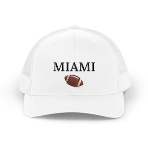 May include: White trucker cap with a mesh back and a curved peak. The word "MIAMI" is printed in black above a brown American football graphic. A casual accessory for sports fans.