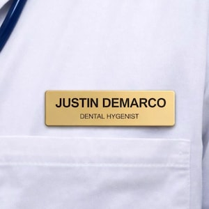 Pode incluir: Close-up de um jaleco branco com uma placa dourada que diz "JUSTIN DEMARCO, DENTAL HYGIENIST". Um estetoscópio azul é visível. O jaleco tem um bolso no lado esquerdo.