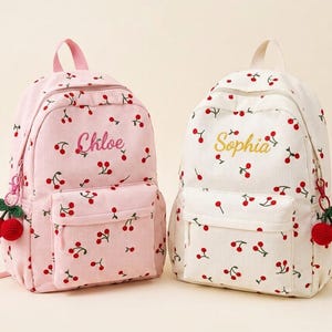 Pode incluir: Duas mochilas de veludo cotelê com estampa de cerejas. A mochila da esquerda é rosa claro com o nome Chloe bordado em rosa. A mochila da direita é off-white com o nome Sophia bordado em amarelo. Ambas possuem um puxador de zíper em formato de cereja.