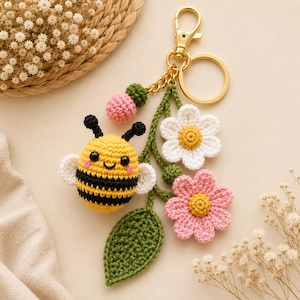 May include: A crochet keyring with a yellow and black bee, white wings, a green leaf, and two flowers, one white and one pink. The keyring has a gold-coloured clasp and ring.