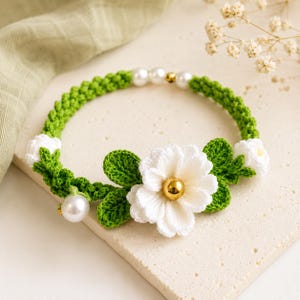 May include: A crochet necklace featuring a vibrant green braided band adorned with white crocheted flowers, green leaves, and smooth white pearls. A large, central white flower is accented with a polished gold-tone bead at its center.