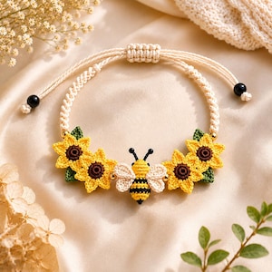 Könnte beinhalten: Ein handgefertigtes Makramee-Armband mit gehäkelten Sonnenblumen und einer Biene. Das Armband hat eine cremefarbene, verstellbare Kordel mit schwarzen Perlen an den Enden. Die Sonnenblumen sind gelb mit braunen Zentren und grünen Blättern.