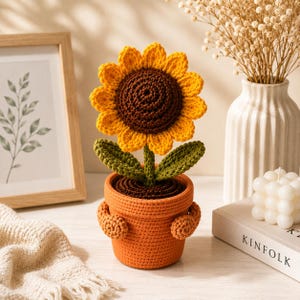 Könnte beinhalten: Eine gehäkelte Sonnenblume mit dunkelbrauner Mitte und leuchtend gelben Blütenblättern, befestigt an einem grünen Stiel mit zwei Blättern. Die Blume steht in einem strukturierten orangefarbenen Topf mit zwei kleinen, runden Häkelarmen.