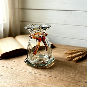 May include: A clear glass vase with a ruffled, flared rim and a rounded body. The vase features a decorative amber-colored glass ribbon tied around the neck in a bow shape, with two long, dangling ends extending down the front of the glass vessel.