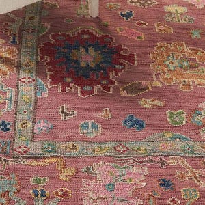May include: A detailed close-up of a pink rug with an intricate pattern. The rug features a central design with red, blue, and yellow accents, surrounded by a border with a fringe. The rug is placed under a white chair leg.