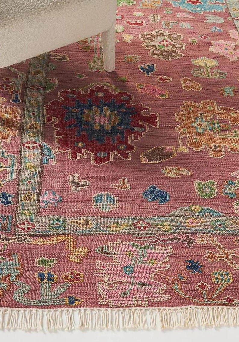 May include: A detailed close-up of a pink rug with an intricate pattern. The rug features a central design with red, blue, and yellow accents, surrounded by a border with a fringe. The rug is placed under a white chair leg.