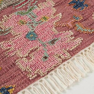 May include: Close-up of a section of a rug with a textured, floral design. The rug features a dusty rose background with pink, blue, and cream-colored floral patterns. The edge has a cream-colored fringe.