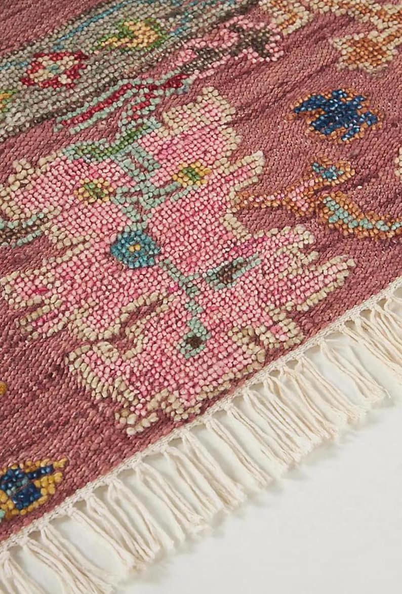May include: Close-up of a section of a rug with a textured, floral design. The rug features a dusty rose background with pink, blue, and cream-colored floral patterns. The edge has a cream-colored fringe.