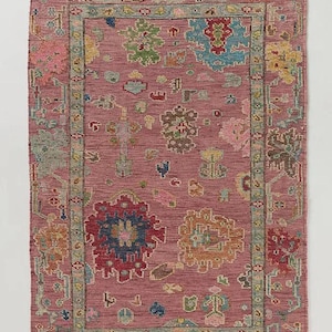 May include: A rectangular rug with a dusty rose background and a detailed floral pattern. The rug features a border and central design with various colors including red, blue, green, and yellow. The edges have a fringed trim.