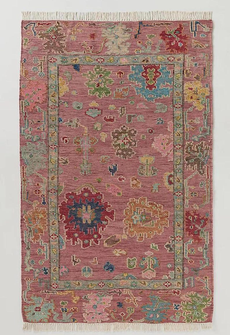 May include: A rectangular rug with a dusty rose background and a detailed floral pattern. The rug features a border and central design with various colors including red, blue, green, and yellow. The edges have a fringed trim.
