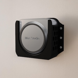 May include: A black wall mount for a Mac Studio computer. The mount has a rectangular shape with rounded corners and a series of ventilation holes on the side. The Mac Studio is visible with the text "Mac Studio" on the front.