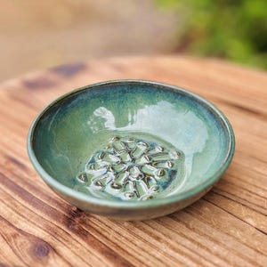 May include: A handmade ceramic bowl with a glossy, teal-green glaze. The interior features a textured, raised design. The bowl is set on a wooden surface, showcasing its unique craftsmanship and artistic appeal. The bowl is approximately 6 inches in diameter.