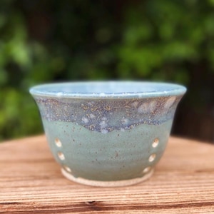May include: A handmade ceramic bowl with a light blue rim and a green body. The bowl has a series of small, evenly spaced holes around the sides. The bowl is sitting on a wooden surface.