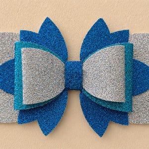 May include: A layered hair bow in shades of blue and silver glitter. The bow features a central teal accent and is designed with multiple layers for a dimensional effect. The bow is a hair accessory.