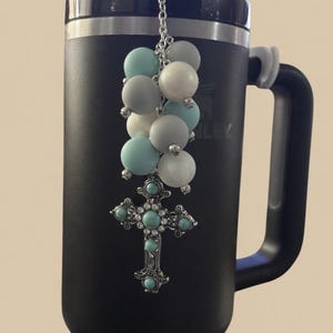 May include: A matte black insulated tumbler with a silver-tone chain charm featuring round beads in shades of white, light blue, and grey, finished with a decorative silver-tone cross pendant inlaid with small turquoise-colored stones and clear crystals.