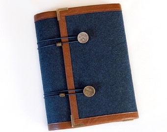 Puede incluir: Una cubierta de cuaderno de fieltro azul oscuro con ribete de cuero marrón y dos detalles en latón. La cubierta tiene dos bandas elásticas negras con cuentas de latón.