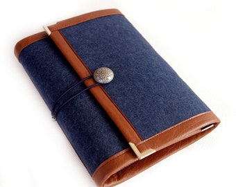 Puede incluir: Un cuaderno de fieltro azul oscuro con ribete de cuero marrón y cierre de botón metálico. El cuaderno tiene un aspecto rústico y vintage.