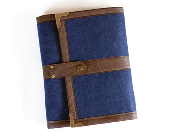 Puede incluir: Una cubierta de cuaderno azul oscuro con detalles en cuero marrón y herrajes de latón. La cubierta tiene una solapa con cierre de botón.