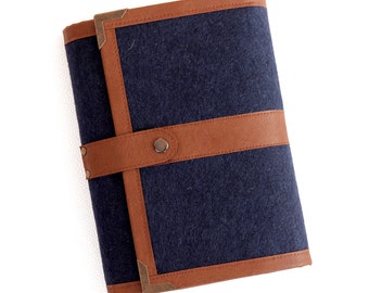 Puede incluir: Un cuaderno de fieltro azul oscuro con ribetes de cuero marrón y detalles en latón. El cuaderno tiene un cierre de solapa y un botón.