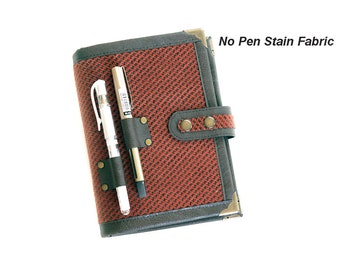 Puede incluir: Un cuaderno de tela rojo y negro con un portaplumas y dos bolígrafos. El cuaderno tiene una solapa con dos botones de latón y una esquina de metal. El texto "No Pen Stain Fabric" está impreso en el lado derecho de la imagen.