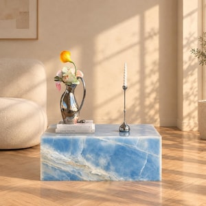 Peut inclure: Une table basse rectangulaire en marbre bleu avec des veines blanches. Sur le dessus se trouvent une carafe métallique argentée contenant un lys calla jaune et des fleurs roses, deux livres empilés Kinfolk et Louis Vuitton, et un bougeoir argenté.