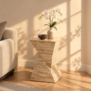 May include: A beige travertine side table with a geometric, twisted hourglass shape. On top sits a light-colored ceramic vase containing white orchid flowers and a small, dark, round bowl. The table features a natural, porous stone texture.