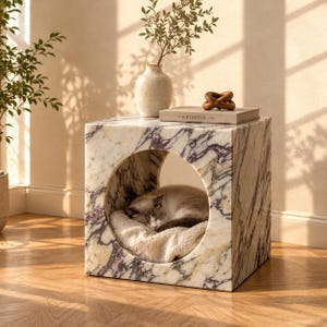 May include: A cube-shaped pet bed made of white and purple veined marble with a large circular opening in the front. Inside the hollow centre, a light-coloured cat is sleeping on a soft, textured cream-coloured cushion. The structure is solid.