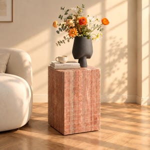 May include: A square side table constructed from vertical pink stone tiles. On top sits a matte black vase filled with orange and white flowers, a small cream-coloured ceramic mug, and a book with the word Kinfolk printed on the spine.