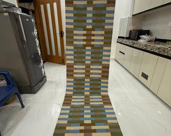 May include: A long, rectangular runner rug featuring a repeating geometric pattern of horizontal and vertical blocks in shades of light blue, olive green, tan, and dark brown, arranged in a woven-style grid across the entire surface.