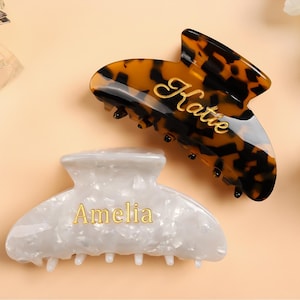 May include: Two personalized hair claw clips. One is tortoiseshell with "Katie" in gold script, the other is white with a pearlescent finish and "Amelia" in gold script. Both are on a light peach background.