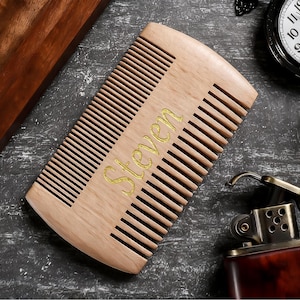 Könnte beinhalten: Ein hellfarbiger Holzkamm mit dem Namen "Steven" in goldener Schrift. Der Kamm hat eng stehende Zähne und ist rechteckig. Ein Feuerzeug im Vintage-Stil und eine Taschenuhr sind ebenfalls im Bild.