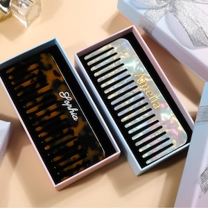 May include: Two personalized combs in gift boxes. One comb has a tortoiseshell pattern with the name "Sophia" in script. The other comb is pearlescent with the name "Amelia" in gold script. The boxes are pastel colors with silver ribbon.