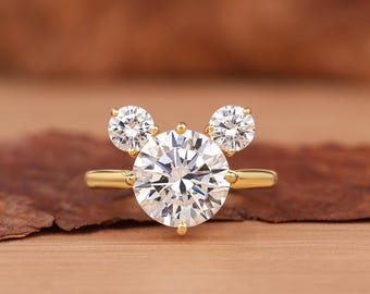 May include: A gold ring featuring a large, round, clear gemstone flanked by two smaller, round gemstones, creating a Mickey Mouse-inspired design. The ring is set against a brown background.