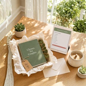 May include: A sage green spiral-bound notebook titled Chores for Couples with a small brown tag labelled For Us with a heart symbol. A tablet displays a digital chore checklist, accompanied by two small potted succulents and a blank envelope.