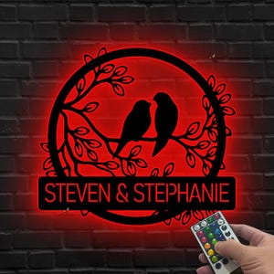 Könnte beinhalten: Schwarze Metall-Wandkunst mit zwei Vögeln auf einem Ast in einem Kreis. Die Namen "STEVEN & STEPHANIE" sind zu sehen. Die Kunst wird mit roten LEDs beleuchtet. Eine Fernbedienung ist sichtbar.