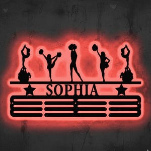 May include: A black metal wall decoration with a red neon glow, depicting cheerleader silhouettes in action. The design includes the name "SOPHIA" and star shapes. The overall design is a decorative piece.
