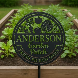 Könnte beinhalten: Schwarzes Metall-Gartenschild mit dem Namen "ANDERSON" und den Worten "Garden Patch FRESH PICKED DAILY" in Grün. Das Schild zeigt Gemüsesilhouetten, darunter Tomaten, Karotten und Erbsen, und ist in einem Gartenbeet platziert.