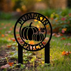 Könnte beinhalten: Schwarzes Metallschild "Pumpkin Patch" mit kreisförmigem Design. Das Schild zeigt zwei Kürbisse und ein Blatt im Kreis, mit den Worten "PUMPKIN PATCH" am Rand. Das Schild ist in den Boden gesteckt.