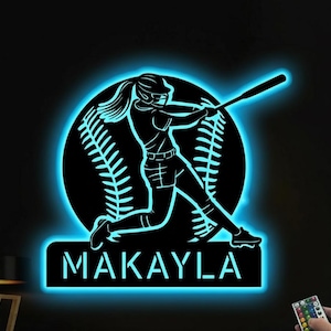 May include: Black baseball-themed wall art with a neon blue glow. The design features a silhouette of a softball player mid-swing, set against a baseball backdrop. The name "MAKAYLA" is displayed below. A remote control is visible.