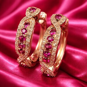 Peut inclure: Deux boucles d'oreilles créoles en or, ornées de pierres précieuses rouges vives et de petits diamants étincelants. Elles sont présentées sur un fond de satin rose, créant une ambiance luxueuse. Les pierres sont d'un rouge profond.